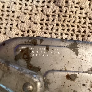 May include: Close-up of a weathered, metallic object with the text "WHEN REVISE WORK REPLACE AT DEALER." The object has a curved shape and appears to be part of a larger mechanism. The background is a cream-colored, textured fabric.