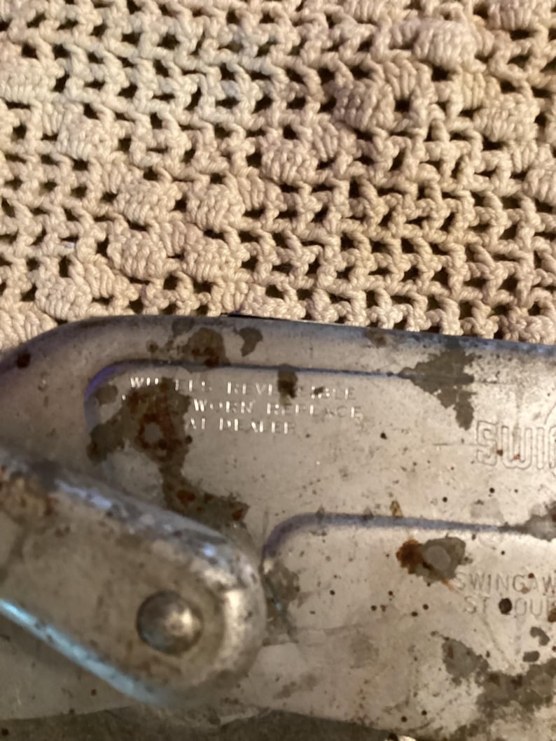 May include: Close-up of a weathered, metallic object with the text "WHEN REVISE WORK REPLACE AT DEALER." The object has a curved shape and appears to be part of a larger mechanism. The background is a cream-colored, textured fabric.
