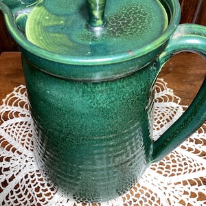 May include: A handcrafted, green ceramic pitcher with a matching lid. The pitcher has a handle and a spout for pouring. The lid has a small knob for easy removal. The pitcher is sitting on a white lace doily.