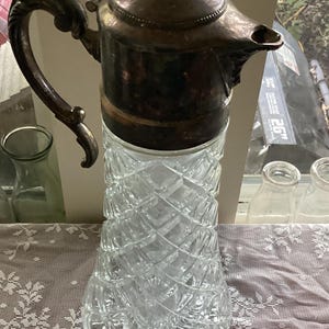 May include: A silver and clear glass pitcher with a decorative handle and a diamond pattern on the glass. The pitcher has a silver lid with a small knob on top.