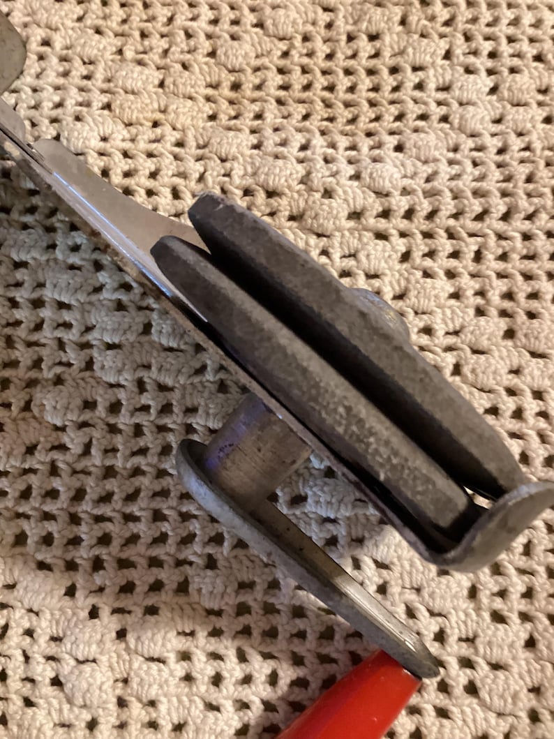 May include: Close-up of a vintage metal tool with a red handle. The tool has a silver blade and a gray, rectangular grip. The tool is designed for a specific purpose, likely related to cutting or shaping materials.