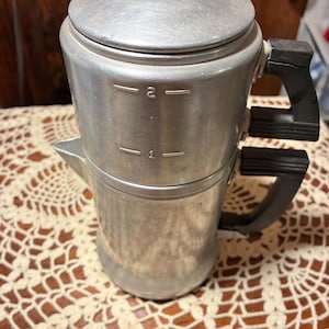 May include: A silver-toned metal coffee percolator featuring a cylindrical body with two black plastic handles on the side. The exterior surface displays embossed measurement markings labeled 1 and 2. It includes a circular lid with a black knob.
