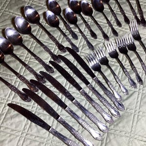 May include: A collection of vintage silverware, including spoons, forks, and knives, arranged on a light green quilted surface. The silverware has ornate, detailed handles and a polished, reflective finish, suggesting a formal dining set.