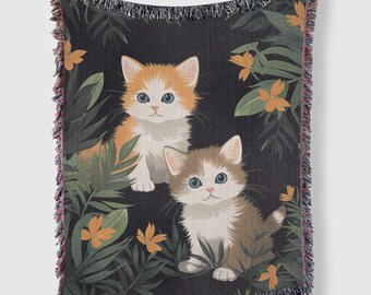 May include: A woven throw blanket with a dark background featuring two kittens with orange and white fur. The kittens are surrounded by green leaves and orange flowers.