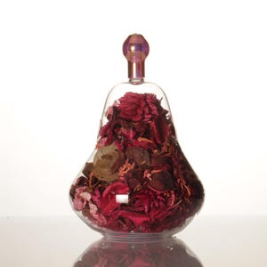 Pode incluir: Uma garrafa de vidro transparente em forma de pêra com uma rolha de vidro roxo cheia de pétalas de flores secas em tons de rosa, vermelho e marrom.