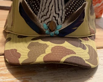 Pode incluir: Um boné de caminhoneiro camuflado com uma malha verde nas costas. O boné é decorado com um grupo de penas em tons de preto, branco e marrom, acentuadas com pedras turquesa.
