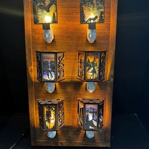 May include: A wooden display featuring six illuminated night lights. Each night light has a unique laser-cut design, including a wolf, a fairy, and other nature scenes. The lights are plugged into a wooden frame, creating a warm, ambient glow.