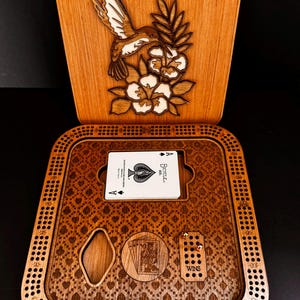 May include: Wooden cribbage board with a hummingbird and floral design on the lid. The board features a playing card holder with a deck of cards, a diamond-shaped cutout, and a section labeled "WINS". The board is made of light brown wood.