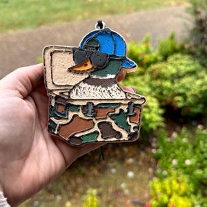 May include: A duck ornament wearing sunglasses and a blue cap, inside a camouflage box. The duck has a green head, a yellow beak, and a white chest. The box is brown, green, and tan. A black string is attached to the top.