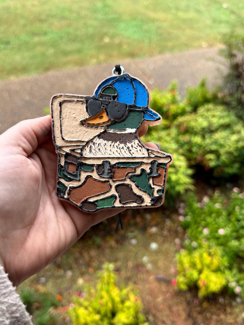 May include: A duck ornament wearing sunglasses and a blue cap, inside a camouflage box. The duck has a green head, a yellow beak, and a white chest. The box is brown, green, and tan. A black string is attached to the top.