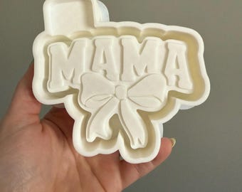 May include: White plastic cookie cutter shaped like the word "MAMA" with a bow underneath.