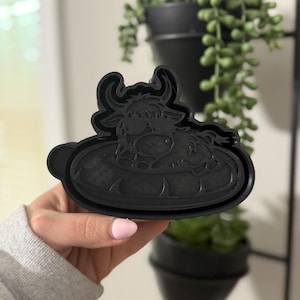 May include: A black plastic cookie cutter in the shape of a Highland cow. The cutter features a detailed design of the cow's face and body, with horns and fur. The cutter is held in a person's hand.