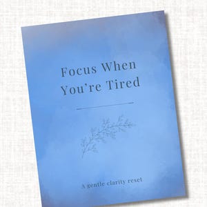 May include: A blue journal with the text "Focus When You're Tired" and "A gentle clarity reset." The cover features a floral illustration and a horizontal line. The background has a watercolor effect.