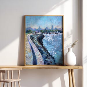 May include: A framed oil painting showing a coastal scene with a pathway, water, and a city skyline. The artwork uses shades of blue and white, with visible brushstrokes. The painting is displayed on a wooden table next to a white vase.