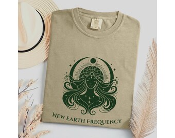 May include: A tan t-shirt featuring a dark green graphic of a stylized figure with long flowing hair, a crescent moon, and sunburst rays. Below the graphic, the text reads NEW EARTH FREQUENCY in a dark green serif font. The shirt has a crew neckline.