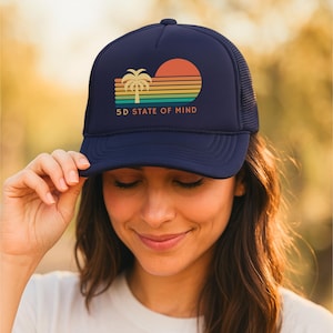 May include: A navy blue trucker hat featuring a graphic print of a palm tree and a setting sun over horizontal stripes in shades of orange, yellow, green, and teal. The text 5D STATE OF MIND is printed in orange below the graphic design.