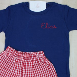 May include: A navy blue short-sleeved t-shirt with the name "Elias" embroidered in red on the front, paired with red and white gingham shorts.