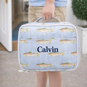 May include: A blue and white striped lunch bag featuring a pattern of fish. The bag has a white zipper and handle. The name "Calvin" is printed in navy blue on the front of the bag.