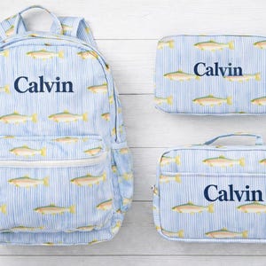 May include: A light blue backpack, lunch bag, and lunch box set. The set features a repeating pattern of fish and vertical stripes. Each item has the name "Calvin" printed in navy blue.