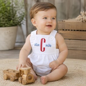 May include: A baby wearing a white romper with a red embroidered letter "C" and blue train designs. The baby is sitting on a rug and playing with a wooden toy train. The romper has button details.