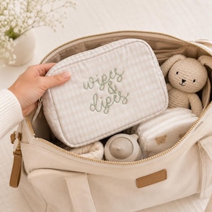 May include: A beige and white gingham zippered pouch with the embroidered text "wipes & dipes" in sage green, placed inside a large canvas tote bag alongside a small crocheted beige bunny toy and a stack of diapers with bear prints.