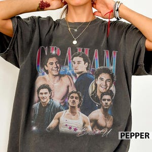 Op de afbeelding: Donkergrijs t-shirt met een collage van afbeeldingen van een man en de naam "GRAHAM" in retrostijl. Het shirt heeft een vintage, versleten uitstraling, met de naam en afbeeldingen in een vervaagde stijl.