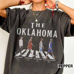 Op de afbeelding: Donkergrijs t-shirt met de witte tekst "THE OKLAHOMA". Daaronder lopen vier figuren over een zebrapad. De namen van de figuren staan eronder afgedrukt. Het woord "PEPPER" staat rechtsonder.