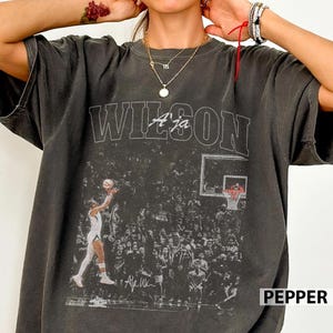 Puede incluir: Camiseta gris oscuro con una escena de baloncesto, el nombre "WILSON" y "A'ja" encima de la imagen. La camiseta tiene un aspecto vintage, con una multitud en el fondo y la palabra "PEPPER" en la esquina inferior derecha.