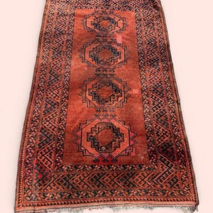 May include: A rectangular, hand-woven rug with a red-orange field and intricate geometric patterns in black and navy blue. The rug features a central design of repeating octagonal shapes and a detailed border. Fringes are visible at the ends.