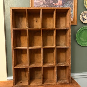 May include: A rectangular wooden storage unit featuring sixteen individual square compartments arranged in a four-by-four grid. The organizer is constructed from light brown wood with visible grain patterns and shows signs of wear.