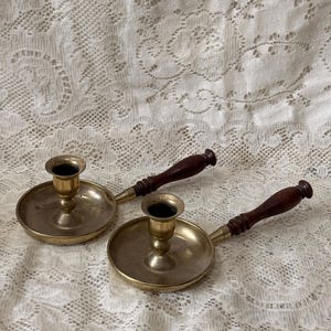 Puede incluir: Dos candelabros de latón con mango, cada uno con una base circular, un receptáculo central para la vela y un mango de madera de color marrón oscuro. Las superficies metálicas muestran un acabado pulido con signos de edad y pátina natural.