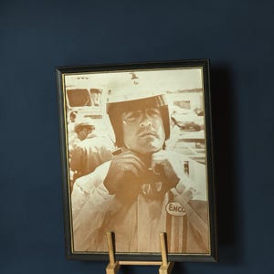 May include: Framed sepia-toned photograph of a race car driver wearing a helmet and racing suit. The driver is adjusting his goggles. The photograph is in a black frame and is displayed on a wooden easel. The ENCO logo is visible on the racing suit.