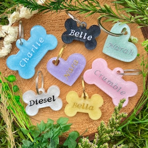 May include: A collection of colorful pet ID tags in various shapes, including bones and hearts. Each tag is a different color, such as blue, black, and pink, and features a pet's name in a contrasting color. The tags are attached to metal rings.