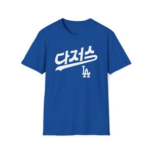 Puede incluir: Una camiseta azul real con un gráfico blanco del texto "LA" en coreano y el logotipo de los Dodgers de Los Ángeles.