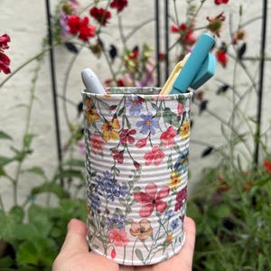 May include: A cylindrical metal container featuring a white background with a colourful watercolour-style floral pattern of red, pink, blue, and yellow wildflowers. The container holds a grey pen and a light blue pen inside its open top.