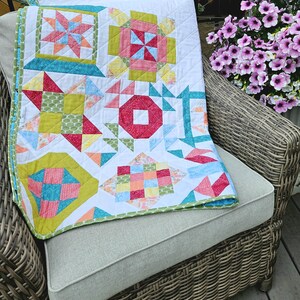 May include: A vibrant patchwork quilt featuring geometric designs in red, pink, blue, green, and yellow. The quilt is displayed on a wicker chair, highlighting its detailed construction. The quilt's edges are finished with a green and white patterned border, adding a decorative touch.