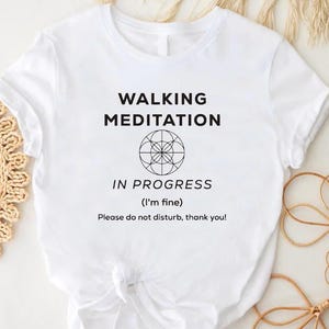 May include: White t-shirt with the text "WALKING MEDITATION IN PROGRESS (I'm fine) Please do not disturb, thank you!" in black. The shirt features a round graphic and is knotted at the bottom.