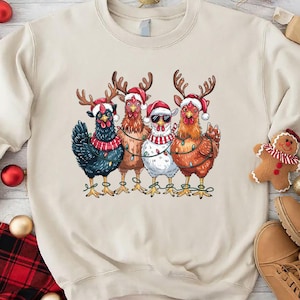 May include: A cream-colored sweatshirt featuring a graphic of five chickens dressed for Christmas. Each chicken wears a Santa hat and reindeer antlers. One chicken wears sunglasses. The chickens are strung with Christmas lights.