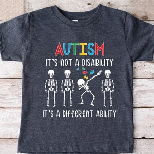 May include: Dark gray t-shirt with the word "AUTISM" in colorful letters. Below the word are the words "IT'S NOT A DISABILITY IT'S A DIFFERENT ABILITY" and a graphic of skeletons, one dabbing with puzzle pieces.