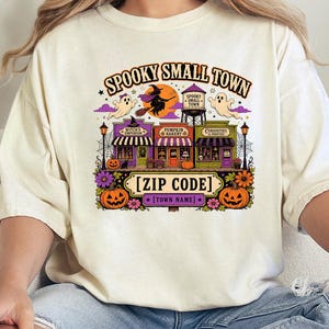 May include: A cream-colored t-shirt featuring a colorful graphic with the text SPOOKY SMALL TOWN, [ZIP CODE], and [TOWN NAME]. The design includes a witch on a broom, ghosts, a water tower, three storefronts, pumpkins, and purple flowers.