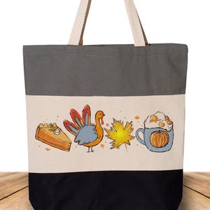 May include: A canvas tote bag with a three-tone design. The top section is gray, the middle is off-white, and the bottom is black. The off-white section features a Thanksgiving-themed illustration with a slice of pie, a turkey, a maple leaf, and a pumpkin spice latte.
