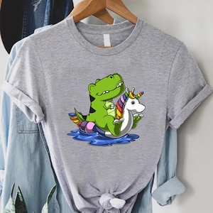 May include: A gray t-shirt featuring a cartoon graphic of a green dinosaur riding a unicorn float in blue water. The unicorn has a rainbow mane and tail. The dinosaur is smiling. The shirt is hanging on a wooden hanger.