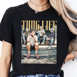 May include: A black t-shirt with the text 'THUG LIFE' in yellow letters. The t-shirt features a photo of four people sitting on a wooden bench. The people are wearing casual clothing and are looking at the camera. The t-shirt is being worn by a person with blue jeans.