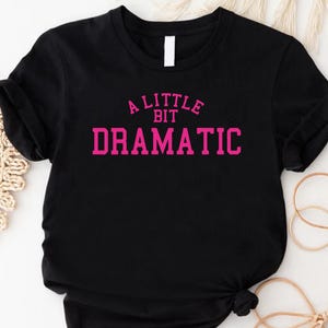 May include: Black t-shirt with the text "A Little Bit Dramatic" in pink, a crew neck, and short sleeves. The shirt is knotted at the bottom. The background includes a woven placemat and decorative elements.