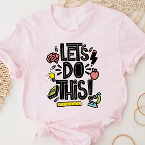 May include: A light pink t-shirt with the phrase "Let's Do This!" in black, with colorful illustrations of a brain, lightbulb, apple, and microscope. The shirt is made of soft material and has short sleeves.