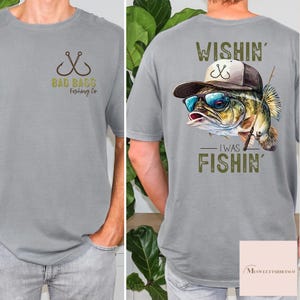 May include: Gray t-shirt with a fishing-themed design. The front features crossed fishing hooks and the text "BAD BASS Fishing Co." The back displays a fish wearing sunglasses and a hat, with the text "WISHIN' I WAS FISHIN'."