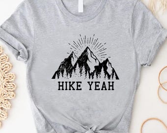 May include: A light gray t-shirt featuring a black graphic of mountain peaks with a sunburst design and the text "HIKE YEAH" in a bold font. The shirt is knotted at the waist.