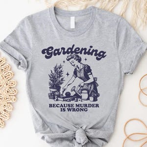 May include: A light gray t-shirt with the words "Gardening Because Murder Is Wrong" in navy blue script. The graphic features a vintage illustration of a woman gardening, with plants and tools.