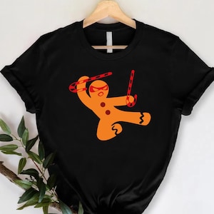 May include: Black t-shirt featuring an orange gingerbread man ninja with a red mask and candy cane weapons. The gingerbread man is in a fighting stance, holding a candy cane in each hand. The shirt is hanging on a wooden hanger.
