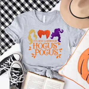 Op de afbeelding: Een grijs T-shirt met een afbeelding van drie heksensilhouetten en de tekst "It's just a bunch of Hocus Pocus".